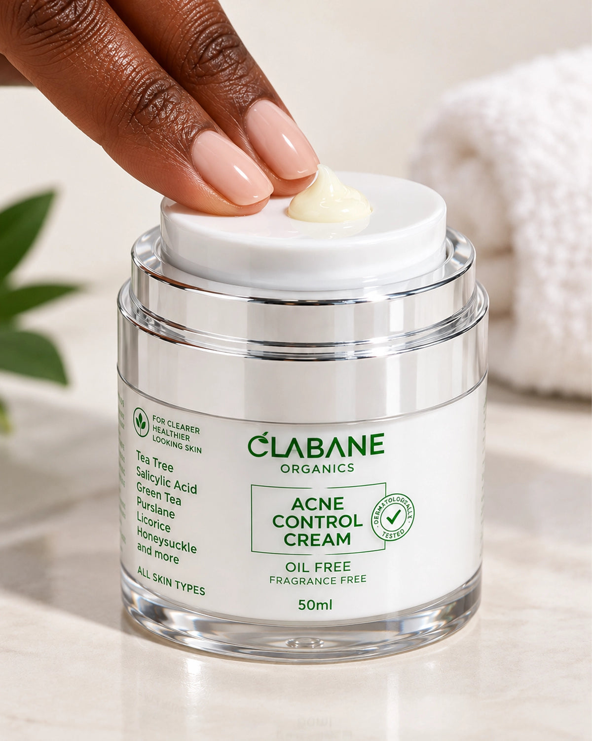 A close-up of a hand with manicured nails lifting the white lid of a Clabane Organics Acne Control Cream jar, with a dollop of pale yellow cream visible on top. The clear glass jar with a white label is set on a light marble surface with a soft, blurred white towel in the background.