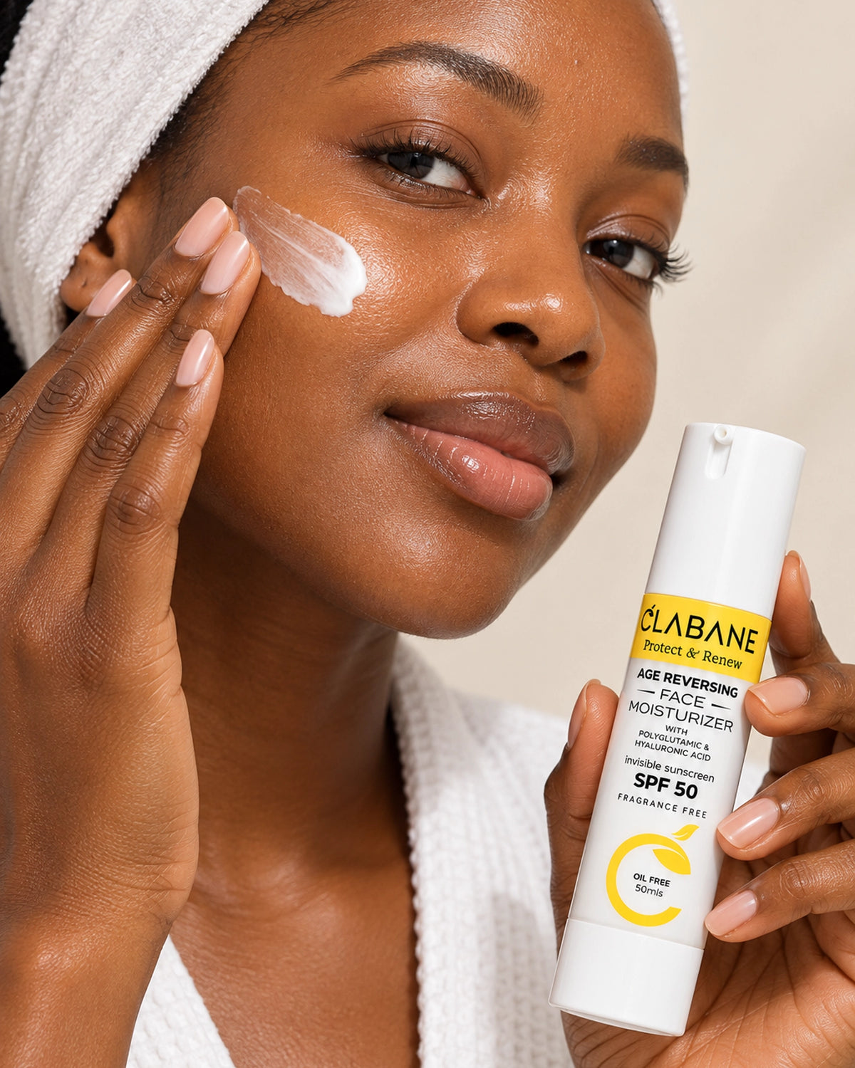 A woman with a white towel on her head applies cream to her cheek while holding a white tube of Clabane Age Reversing Face Moisturizer SPF 50 in one hand.