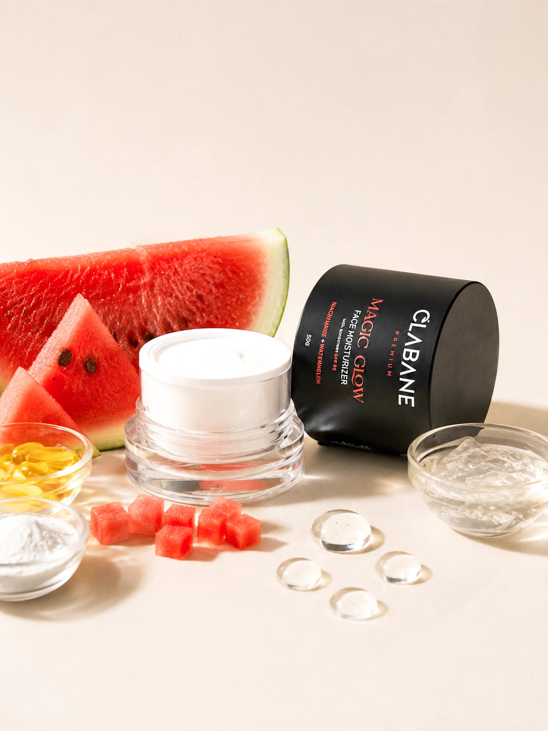 A skincare product set on a warm beige background, featuring a black cylindrical container labeled CLABANE Magic Glow, a white cream jar, watermelon slices, gel cubes, and small glass bowls of ingredients.