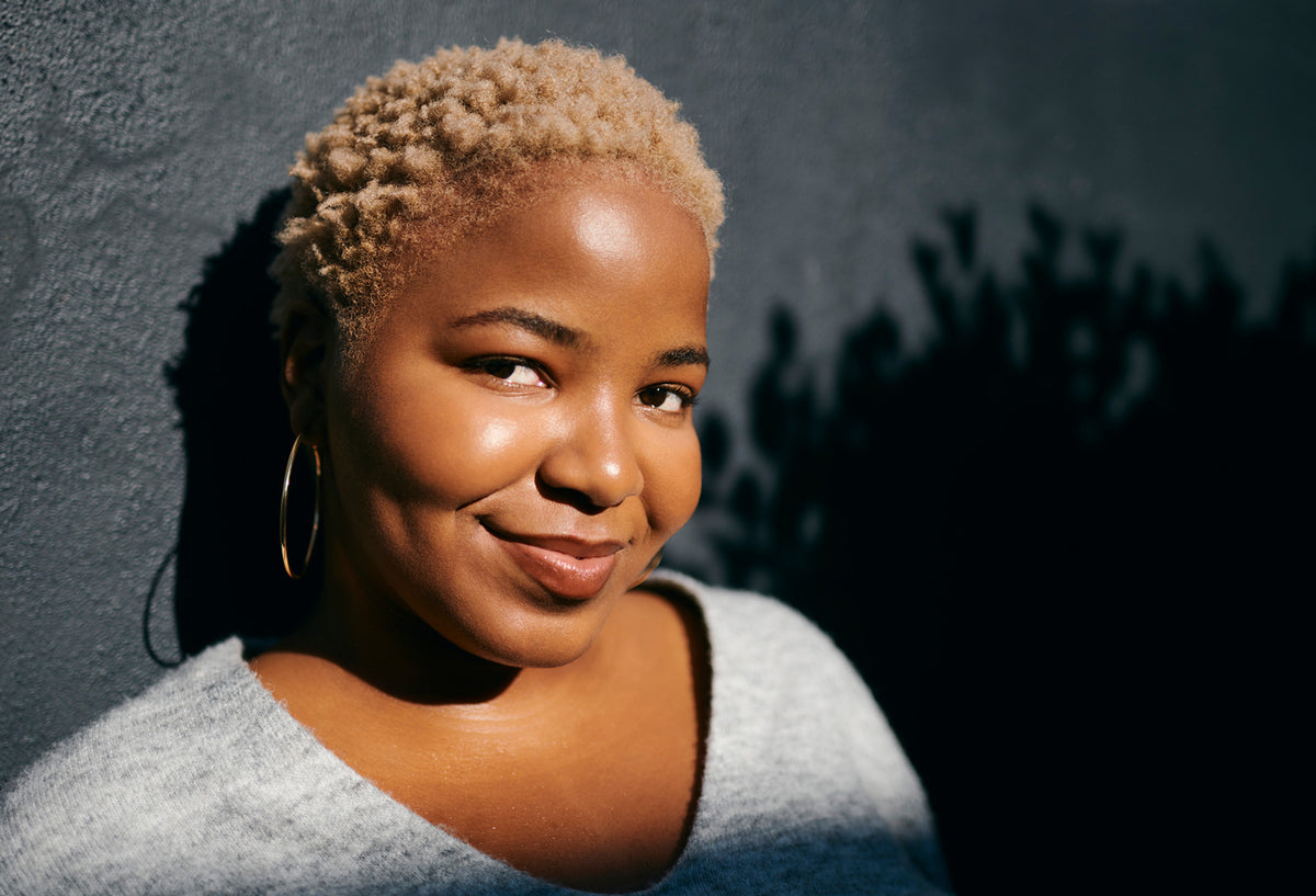 A close-up portrait of a smiling woman with short curly blonde hair, wearing large gold hoop earrings and a light gray top, set against a dark textured wall with dramatic shadows.