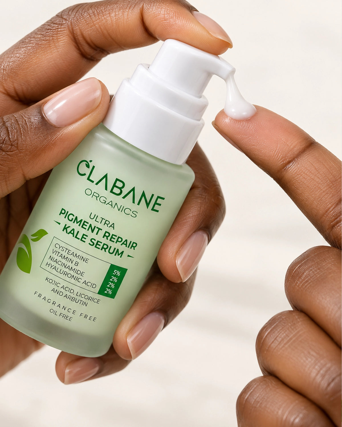 A close-up of a hand holding a bottle of Clabane Organics Ultra Pigment Repair - Kale Serum, with another hand pressing the white pump to dispense a small amount of the serum onto a neutral background.