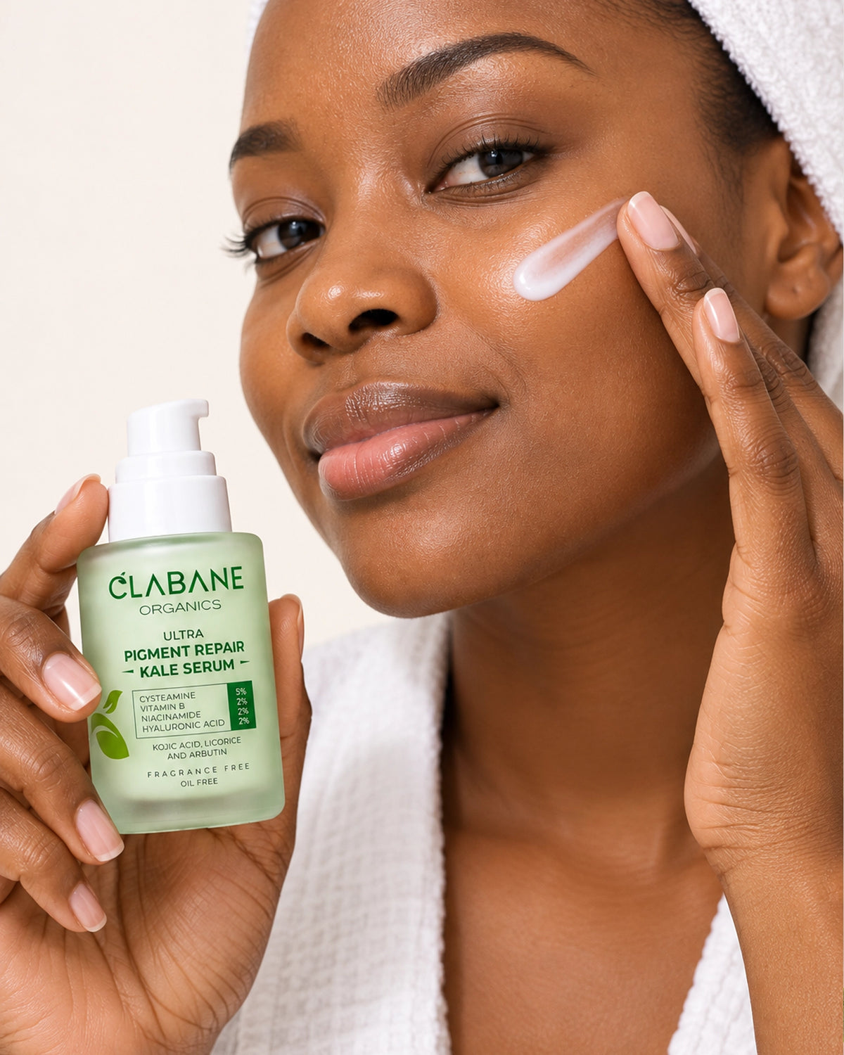A woman with a white towel on her head applies cream to her cheek while holding a bottle of Clabane Organics Ultra Pigment Refining Kale Serum in one hand.