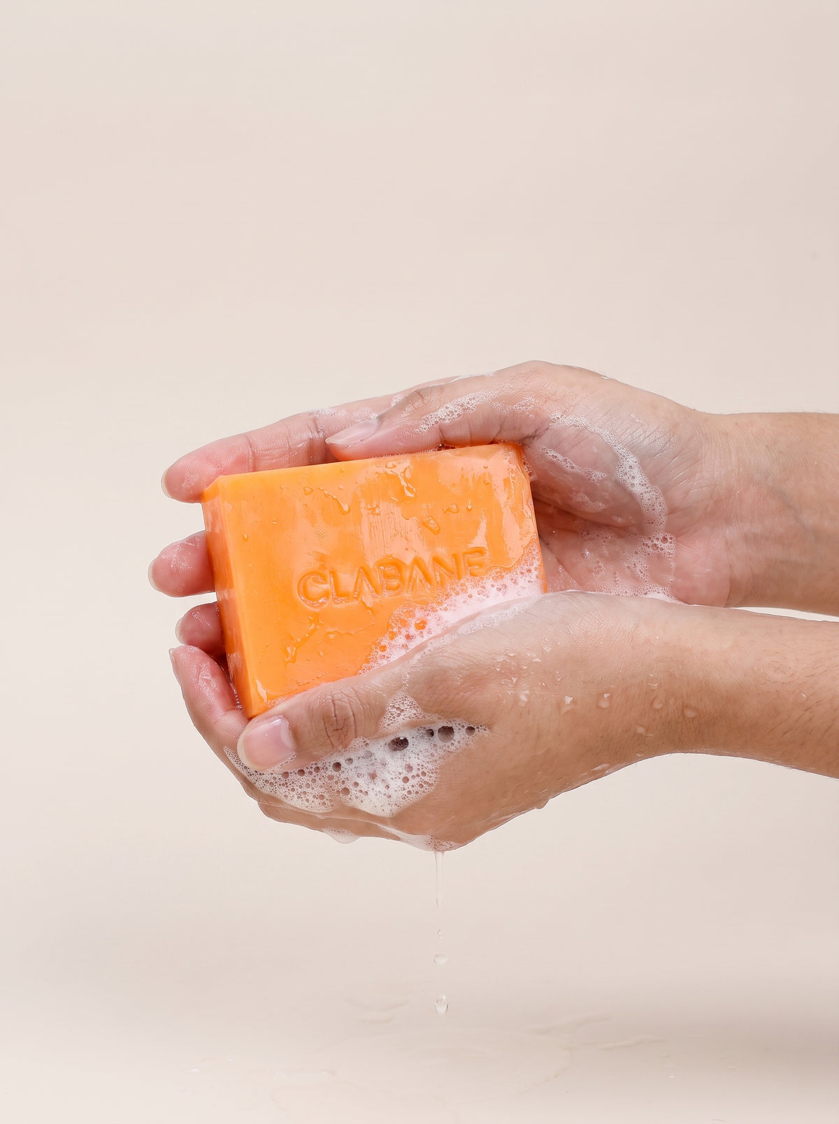 A pair of hands washing a bright orange bar of soap labeled 'CLABANE' with lathered foam, set against a soft beige background.