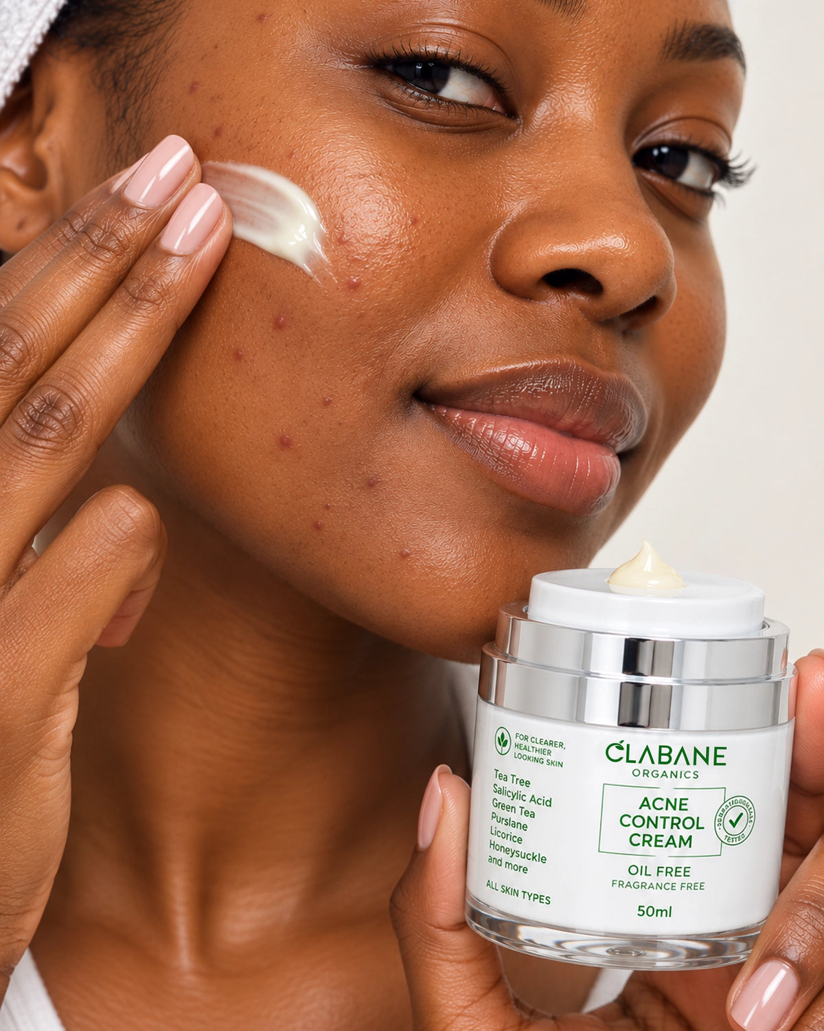 A close-up lifestyle shot of a woman applying cream to her cheek while holding a jar of Clabane Organics Acne Control Cream.