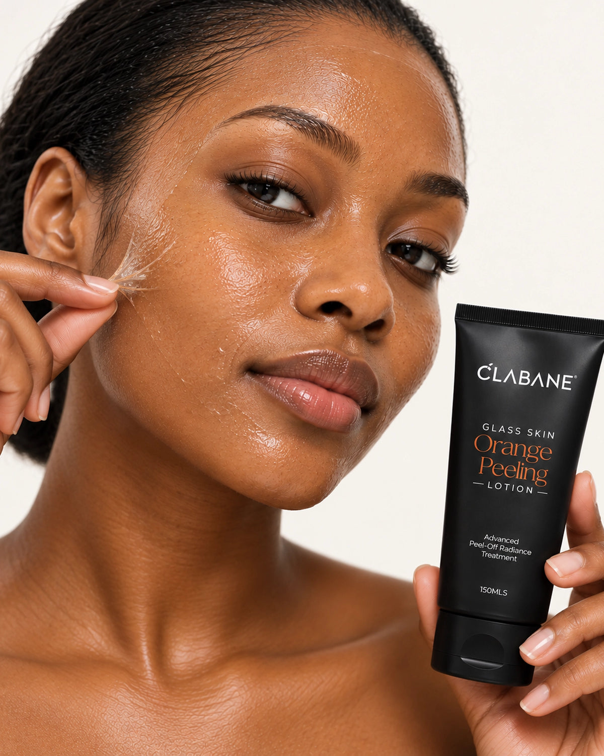 A close-up of a woman with glowing, hydrated skin holding a black tube of Clabane Glass Skin Orange Peeling Lotion next to her cheek where she is applying a clear peel mask.