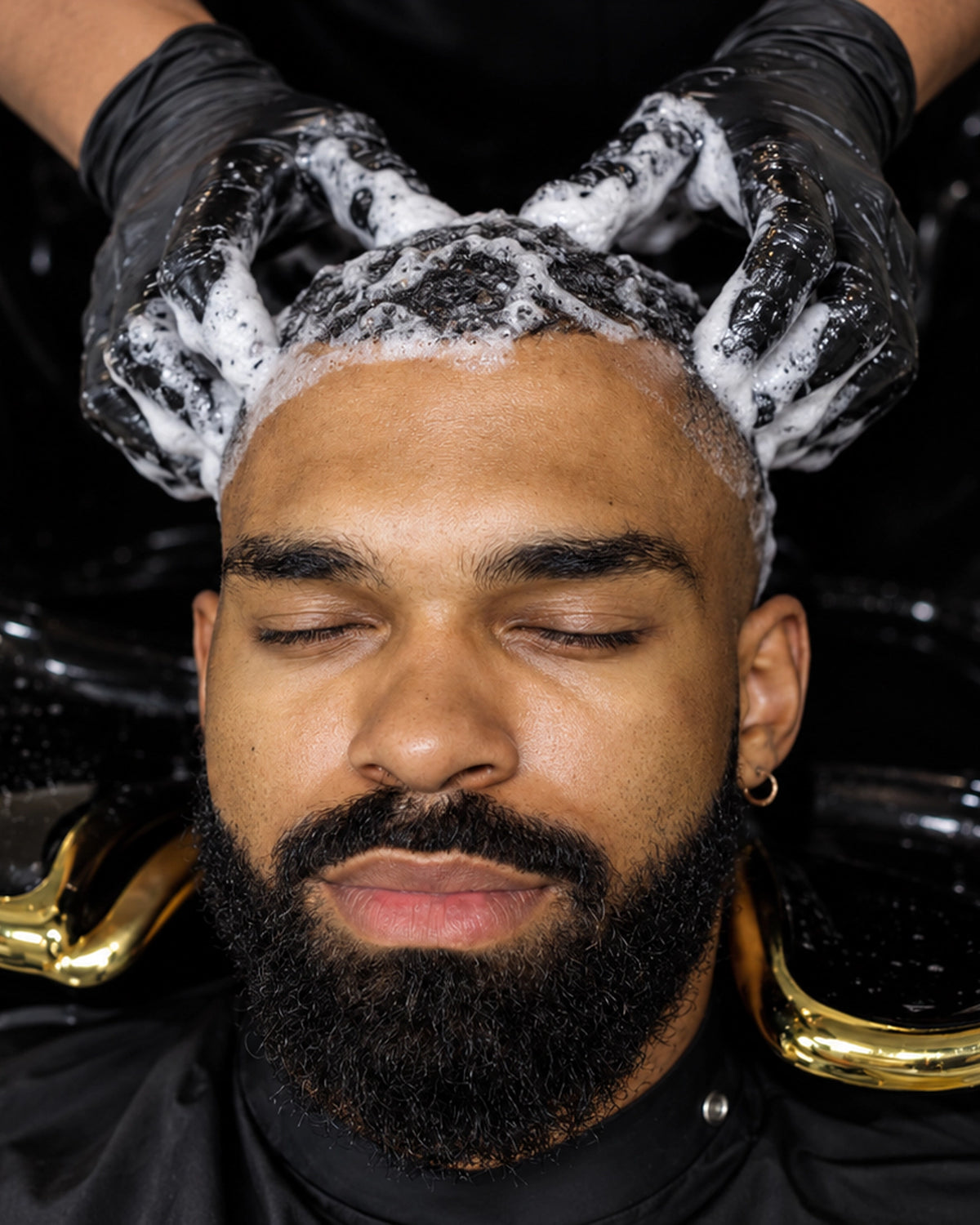 A close-up of a man with a full beard and closed eyes receiving a scalp massage with foamy hair on his head, wearing a black salon cape with gold trim.