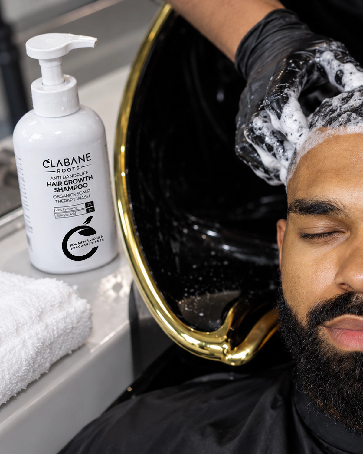 A close-up lifestyle shot of a man receiving a hair wash, with foam lathered on his scalp, next to a black and gold salon sink and a white bottle of Clabane Roots hair growth shampoo on a counter.