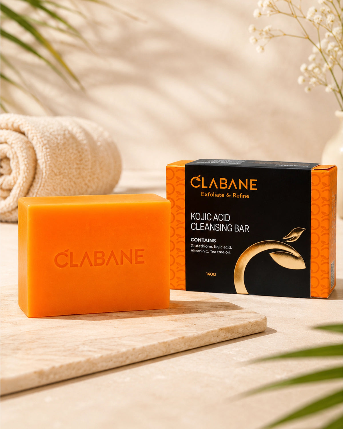 An orange bar of CLABANE Kojic Acid Cleansing Bar sits on a light stone tray next to its black and orange retail box, styled with a rolled towel and soft tropical leaves in a warm, sunlit setting.