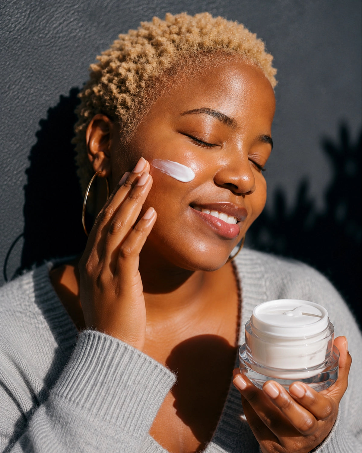 A woman with short blonde hair applies white cream to her cheek while holding an open jar of skincare cream against a dark gray wall.