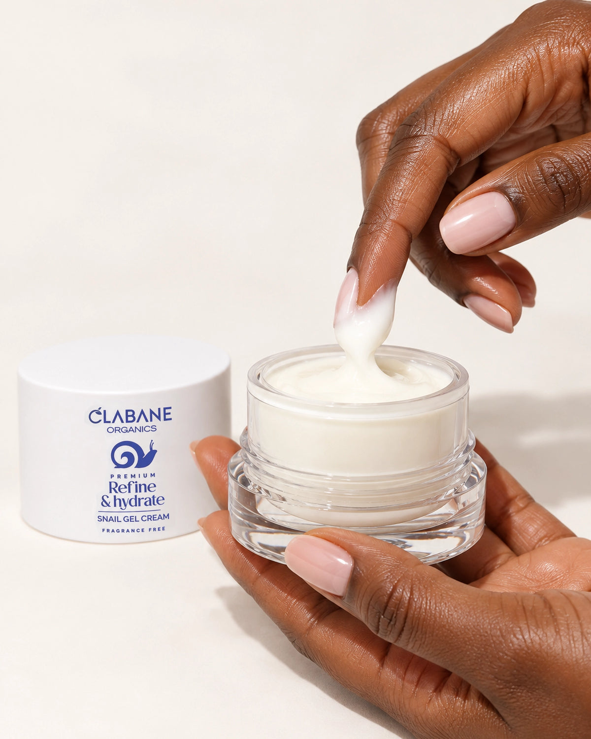 A hand scoops white snail gel cream from a clear jar next to its white lid, which is branded CLABANE ORGANICS. The scene is set against a clean, solid off-white background.