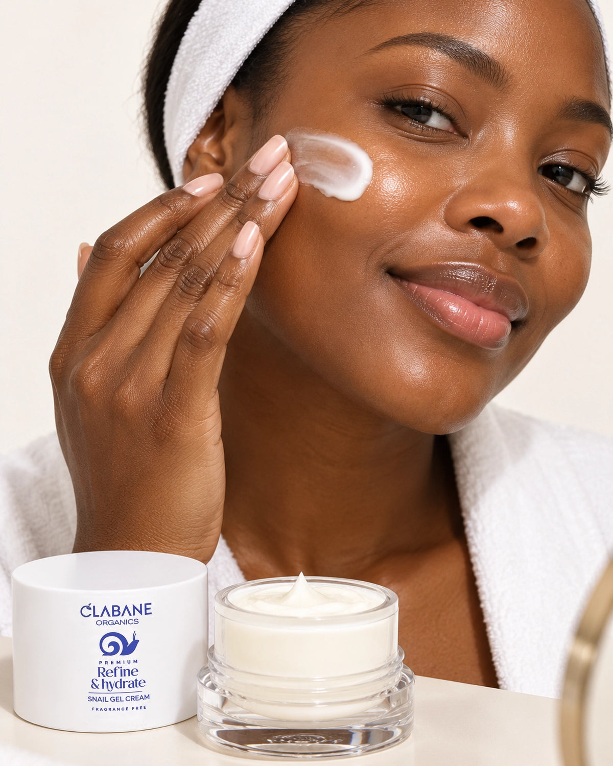 A woman with a white towel headband and robe applies cream to her cheek, with two jars of Clabane Organics Refine & Hydrate facial gel cream in the foreground on a light neutral background.