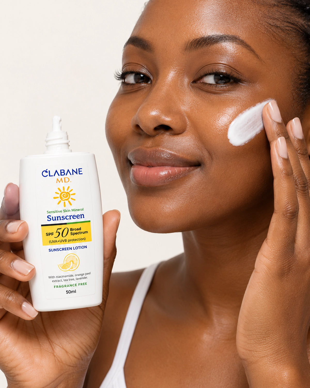 A woman applies white sunscreen lotion to her cheek while holding a bottle of Clabane MD Sunscreen Lotion SPF 50 Broad Spectrum. The bottle is white with blue and yellow branding, set against a clean, light beige background.