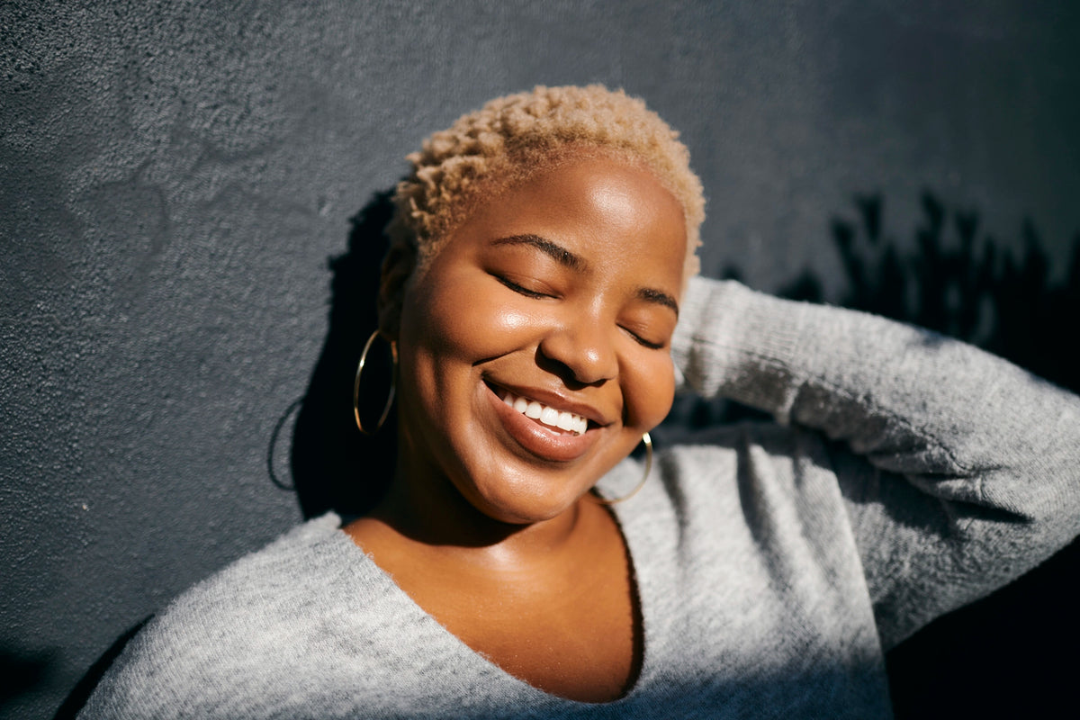 A smiling woman with short blonde hair, wearing a light gray knit sweater and gold hoop earrings, posing against a dark textured wall in dramatic sunlight and shadow.