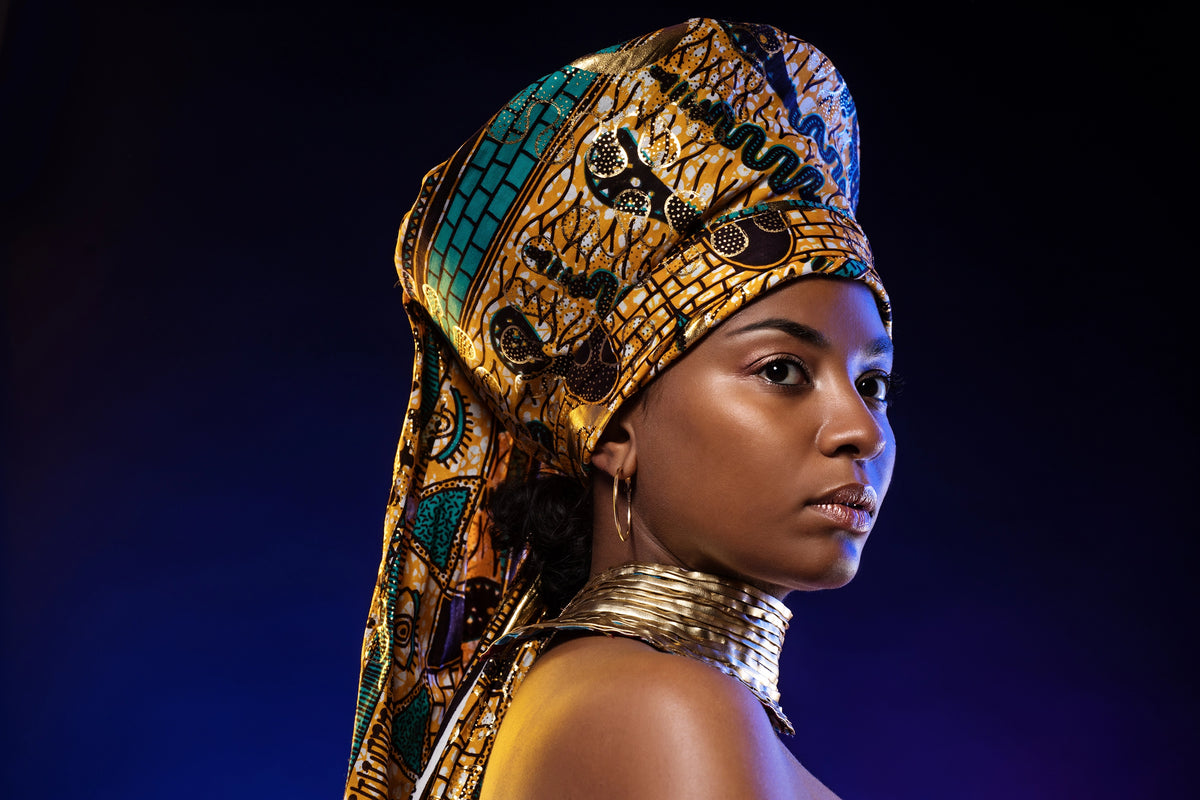 A woman in profile wearing an ornate gold and turquoise headwrap with black detailing and a matching metallic collar, photographed against a dark blue background.