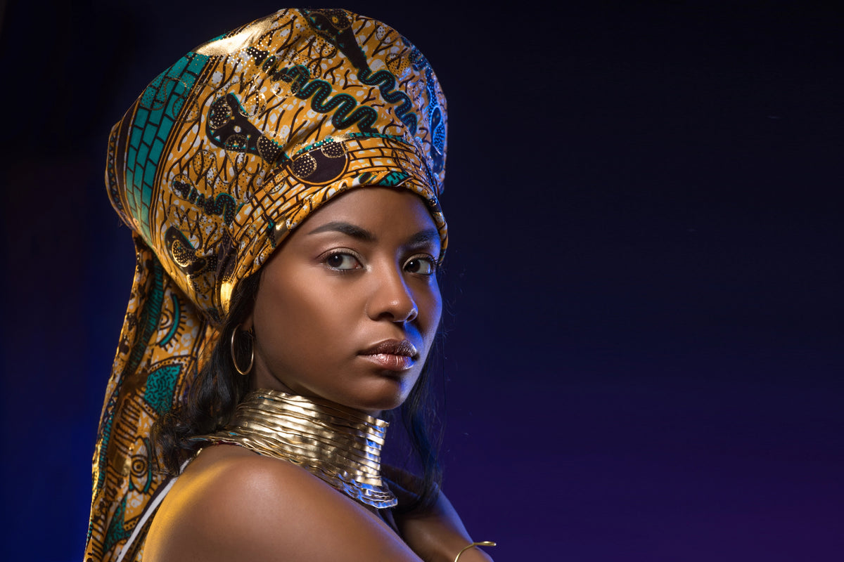 A woman wearing an ornate gold and turquoise patterned headwrap and a chunky gold collar necklace, photographed against a dark blue studio background.