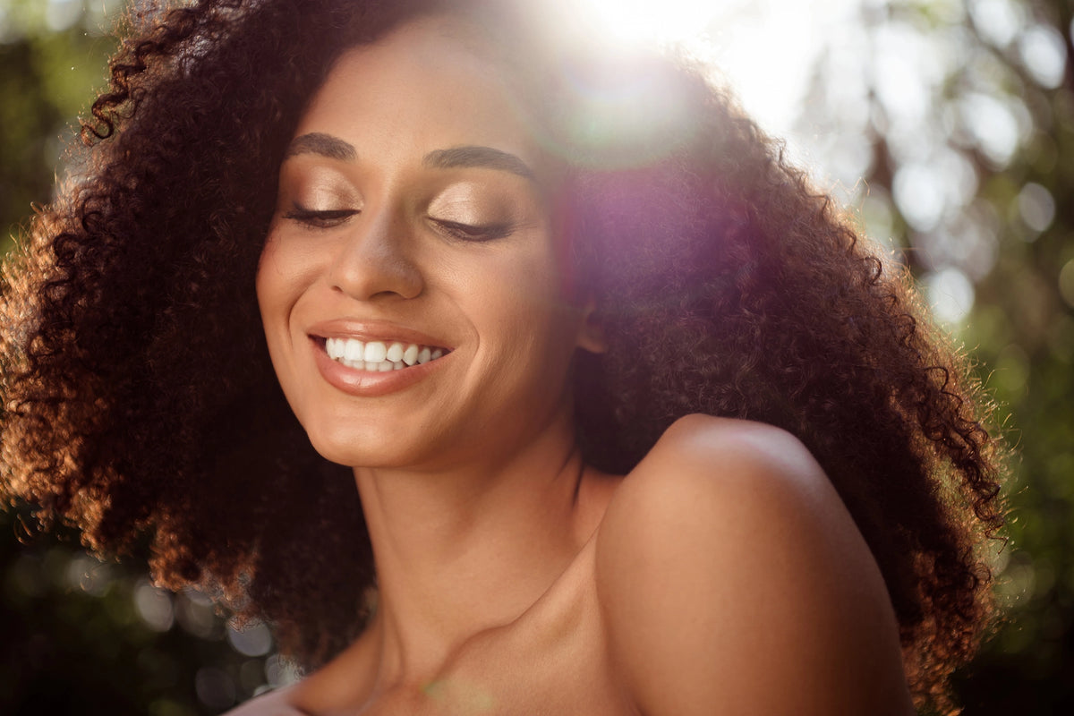 A close-up portrait of a smiling woman with closed eyes and voluminous dark curly hair, set against a bright, sunlit outdoor background with soft green bokeh.