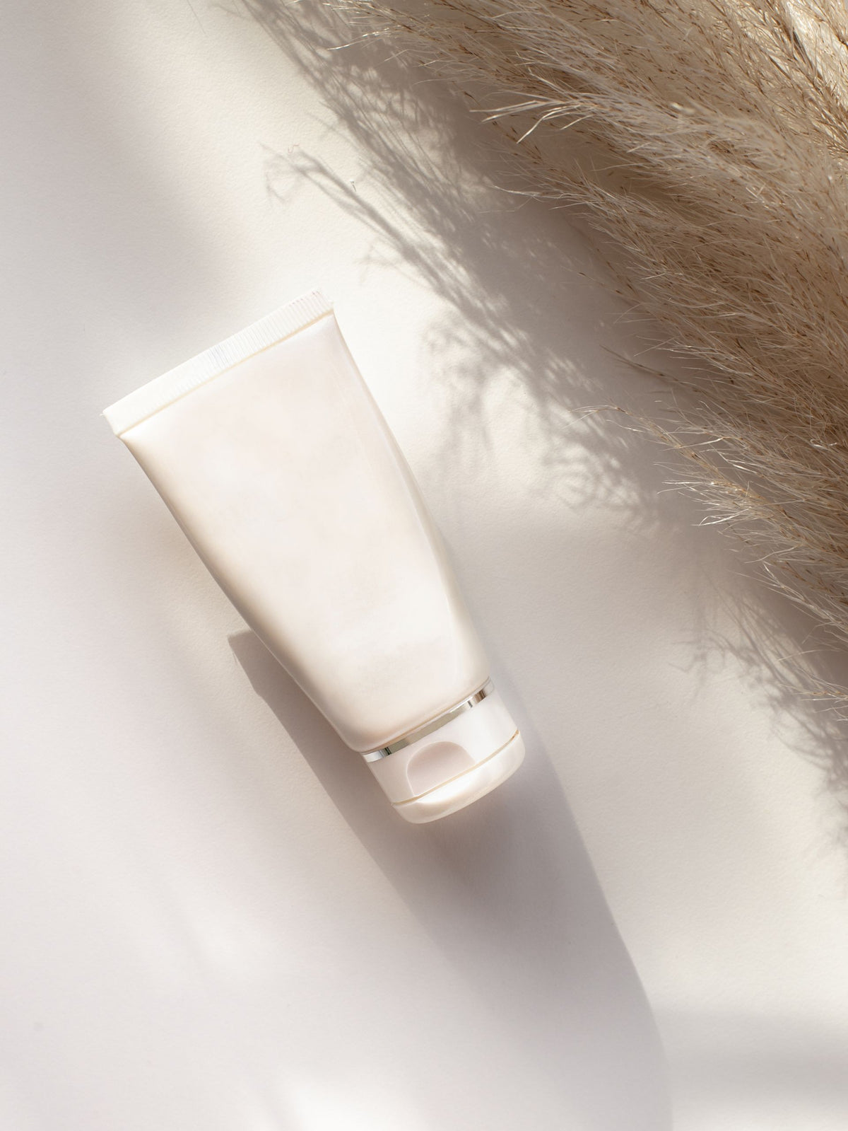 A blank, matte off-white cosmetic squeeze tube with a flip-top cap, photographed diagonally on a white surface with soft shadows and dried pampas grass in the background.