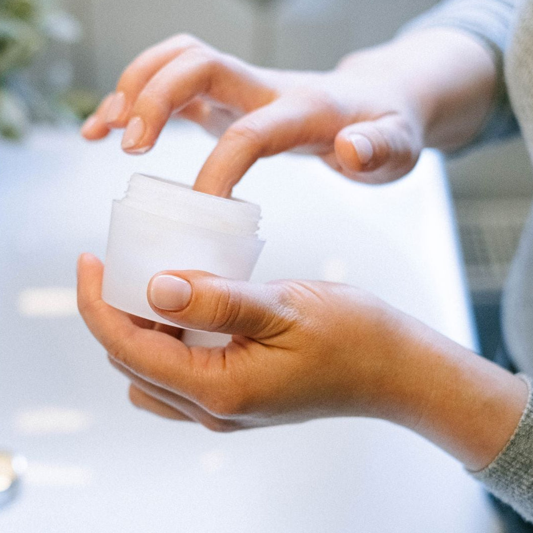 A close-up of a person's hands holding an open white cosmetic jar over a white sink, with a blurred plant and faucet in the background.